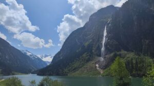 Panoramaansicht des Stillup Stausees in den Alpen, umgeben von beeindruckenden Bergen, einem großen Wasserfall und üppiger Natur unter einem strahlend blauen Himmel.