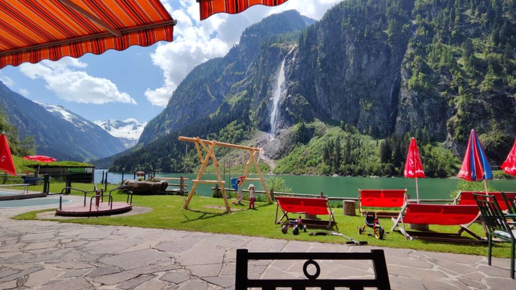 Blick von einer Terrasse mit Sitzgelegenheiten auf den Stillup Stausee, eingerahmt von Bergen und einem großen Wasserfall, während Kinder auf einem Spielplatz am Ufer spielen.