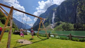 Zwei Kinder schaukeln auf einem Spielplatz am Ufer des Stillup Stausees mit Blick auf einen Wasserfall und die umgebenden Berge in den Zillertaler Alpen.