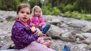 Zwei Kinder sitzen barfuß auf Steinen am Ufer eines Flusses nahe dem Stillup Stausee, eines der Kinder hält einen Snack in der Hand.